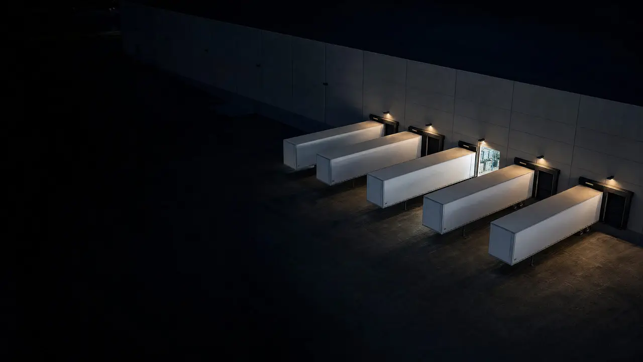 Aerial view of a warehouse dock at night, trailers backed into the doors and one door open on racked pallets.